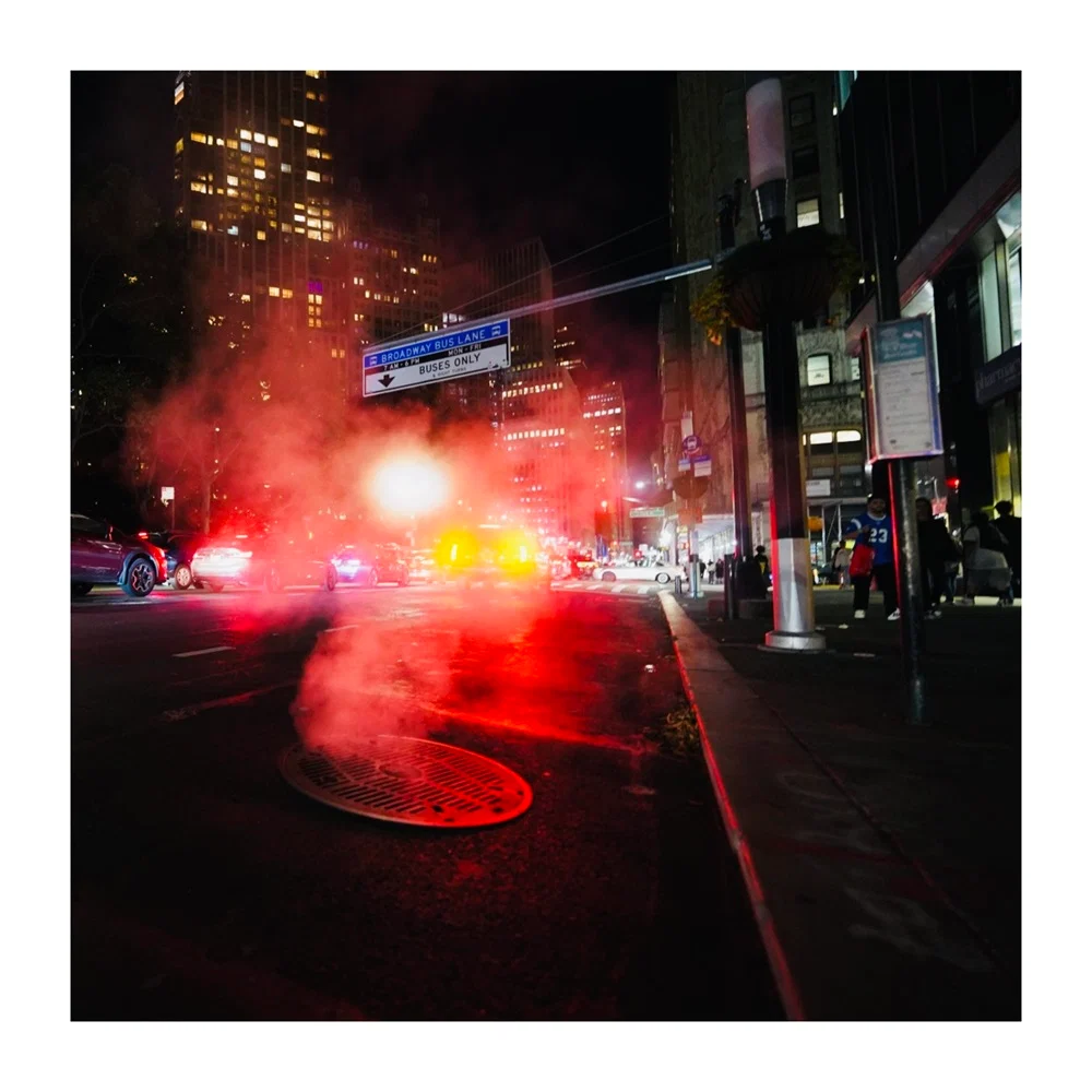 Night shot on a street, car and building lights, smoke coming out of the sewer